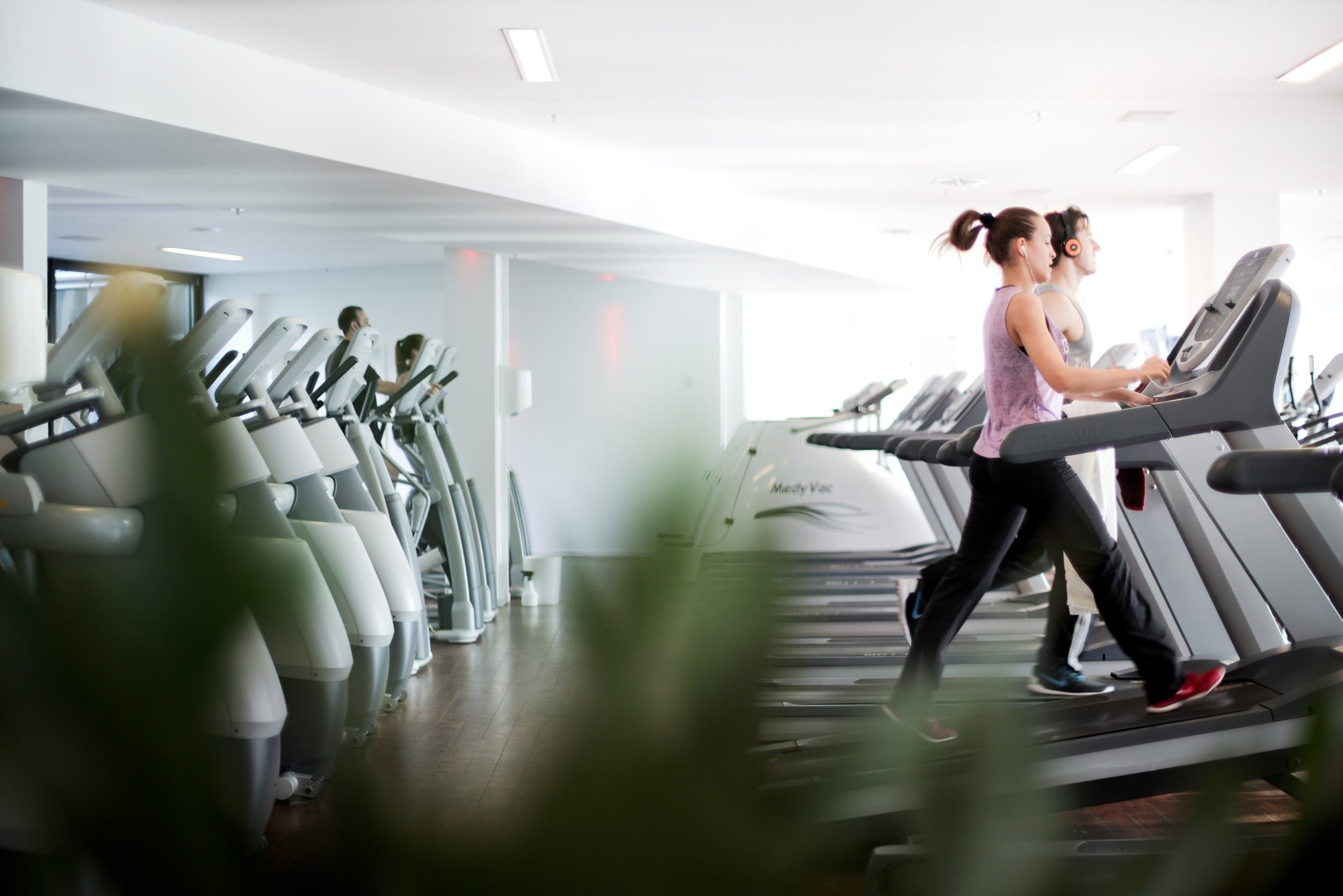 Zusehen  sind zwei Menschen auf dem Laufband. Diese sind versetzt zueinander. Die weiblich gelesene Person im Vordergrund trägt Sportklamotten. eine dunkle Hose und ein rosafarbenes Oberteil. Die Person dahinter wird als männlich gelesen und trägt ebenfalls Sportklamotten.