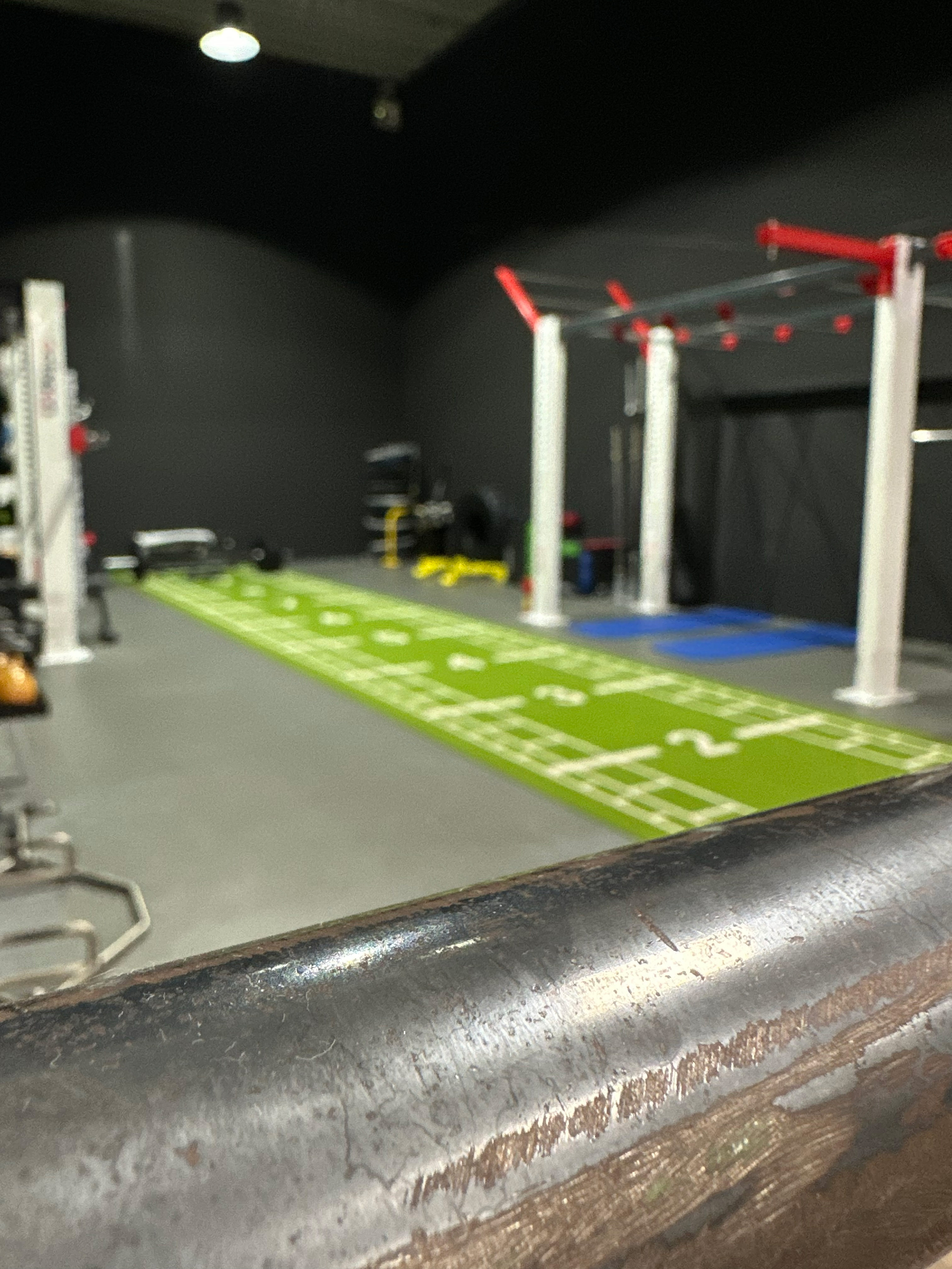 Zu sehen ist eine Nahaufnahme einer Edelstahlstange. Im Hintergrund sieht man einen dunkel gehaltenen Spot und Fitnessbereich, der einen graue Boden und einen grünen Turf beinhaltet