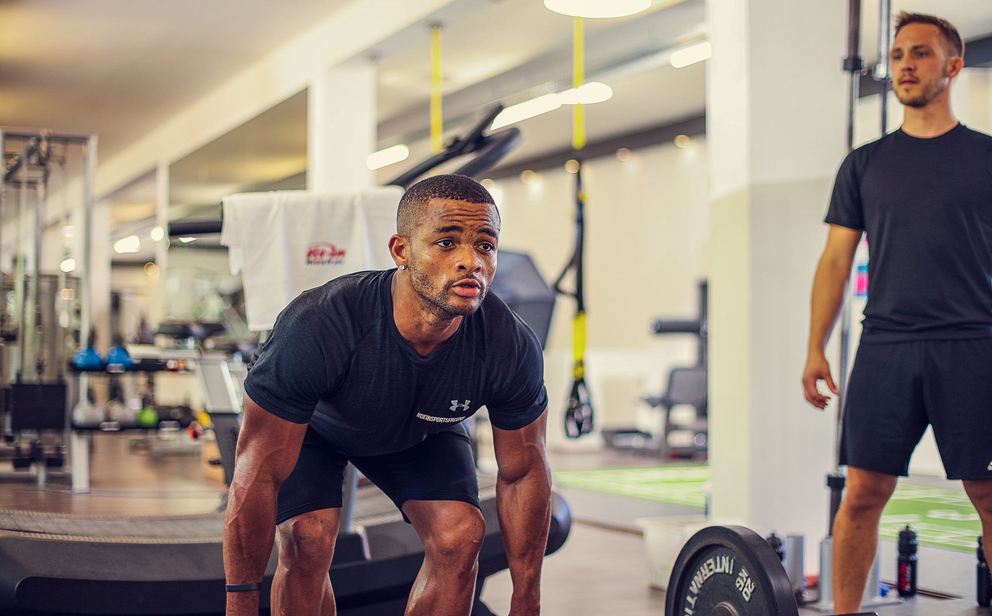 Sportbild. Im Vordergrund beugt sich ein junge tief um eine Kreuzhebe Übung zu machen. Im Hintergrund sieht man das Sportstudi. neben ihm im Anschnitt wird er dabei von einem Trainingspartner beobachtet.