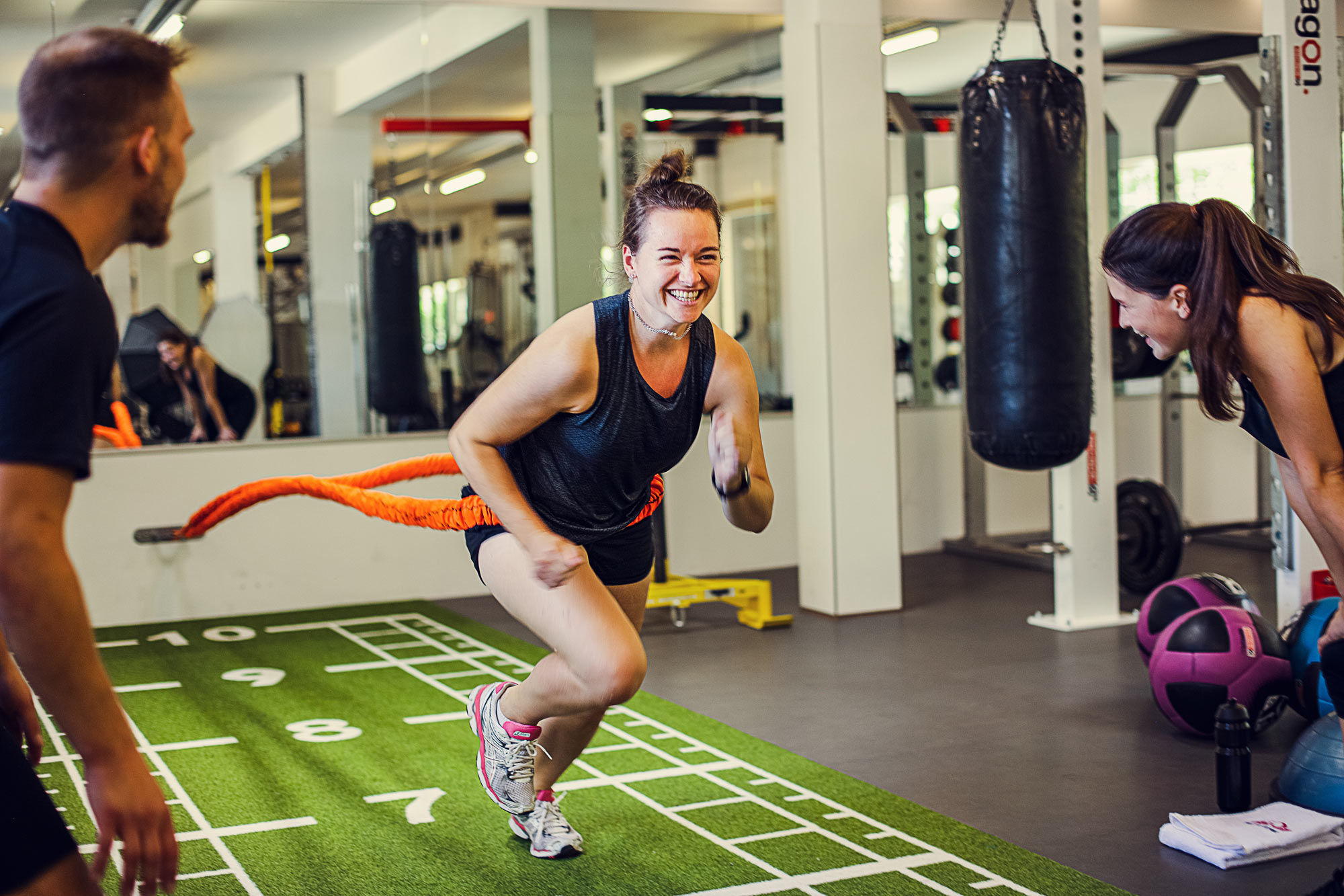 3 Junge Menschen beim Sport. Links aussen steht ein Mann und feuert die Person in der Mitte an. Um die Hüfte der Person in der Mitte ist ein orangenes Bungee Jumpingband welches an der Wand befestigt wurde. rechts aussen steht eine weitere Person um die Person in der Mitte anzufeuern