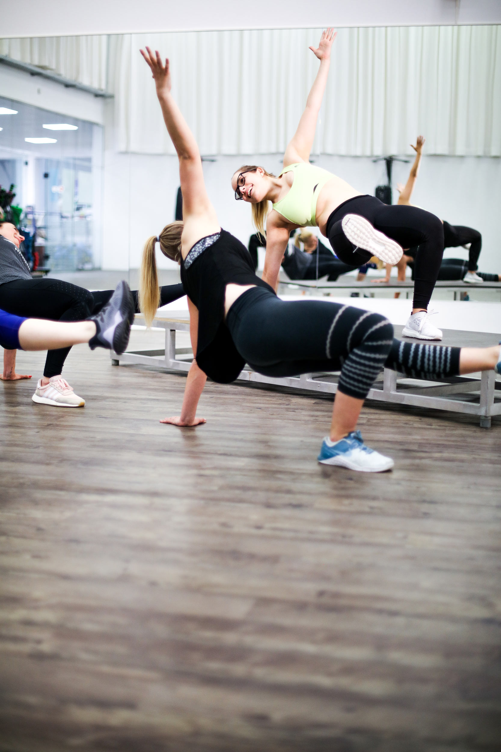 Zu sehen ist eine Sport und Bewegungssituation. Im Vordergrund sind Menschen zu sehen, die eine Art BReakdanceübung machen. Man sieht diese Personen von hinten. Die weiblich gelesene Trainerin macht diese Breakdance Übung vor. Sie ist zu besseren Sichtbarkeit auf einer Erhöhung positioniert,