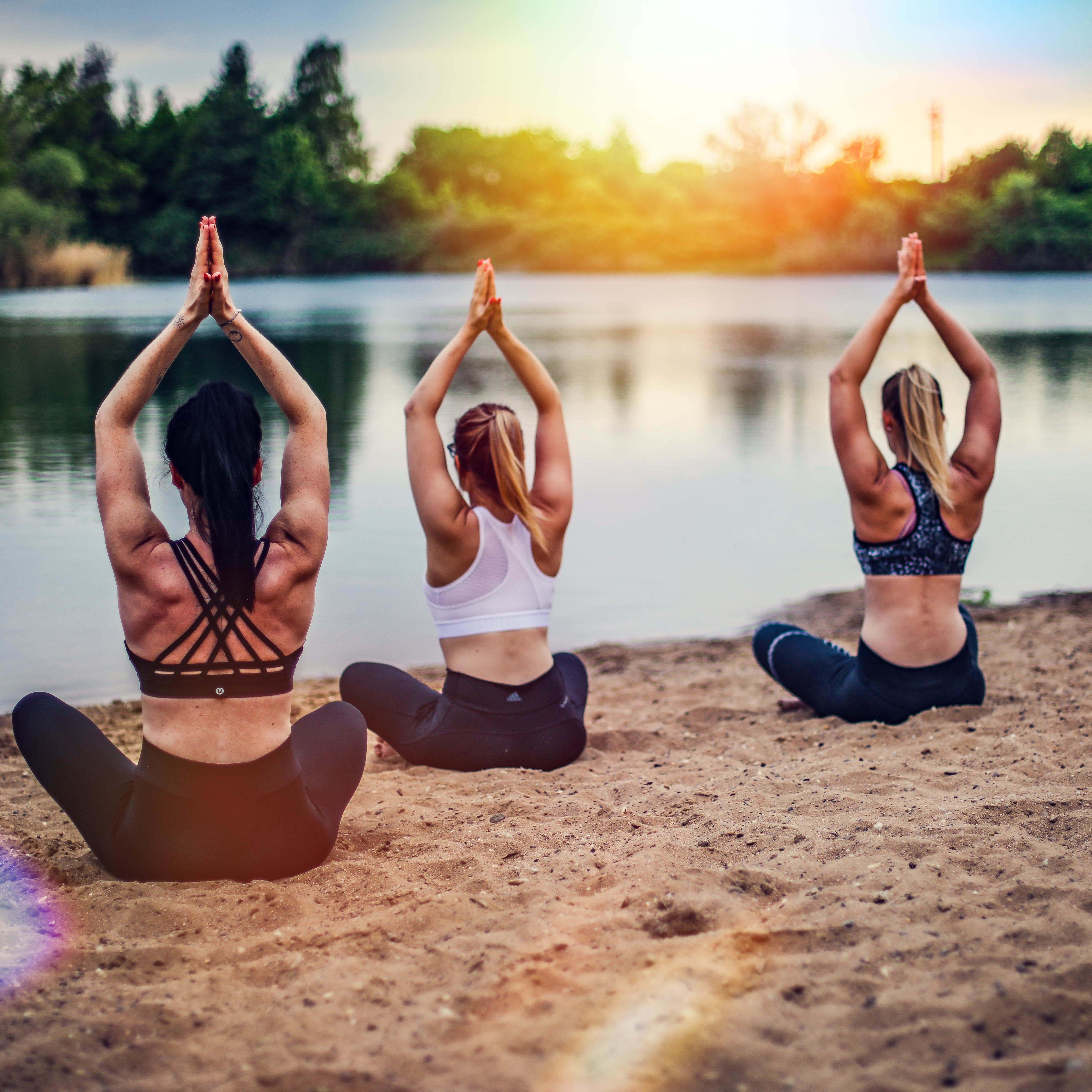 Zu sehen sind drei weiblich gelesene Figuren, die mit gefalteten Händen über den Köpfen am Sandstrand vor einem See sitzen. Alle Personen Sund in leichter Sportbekleidung von hinten zu sehen,
